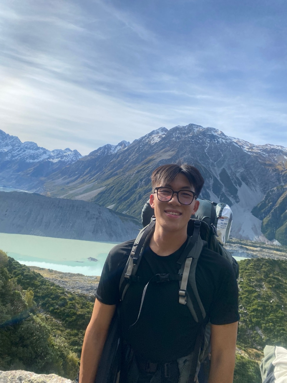 Dom hiking above a glacial lake in New Zealand, with a backpack and snowy mountains behind him