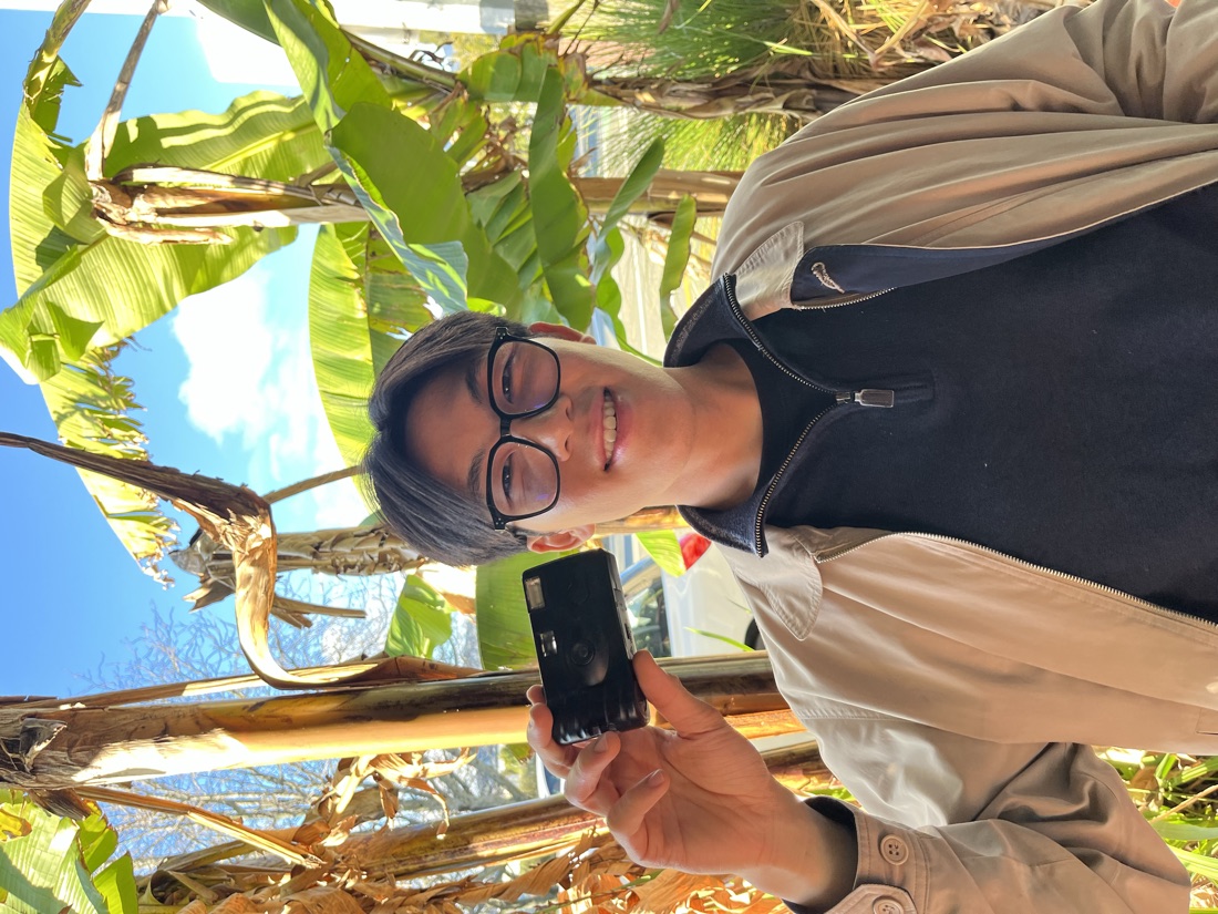 Dom, the person behind Flow Studio Media, holding a film camera in front of banana plants
